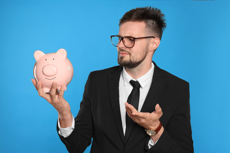 Confused businessman with piggy bank on light blue backgroundの写真素材