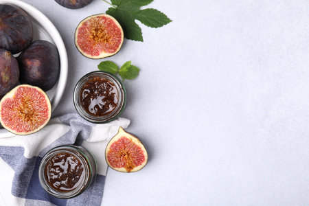 Glass jars of tasty sweet fig jam and fruits on light table, flat lay. Space for text