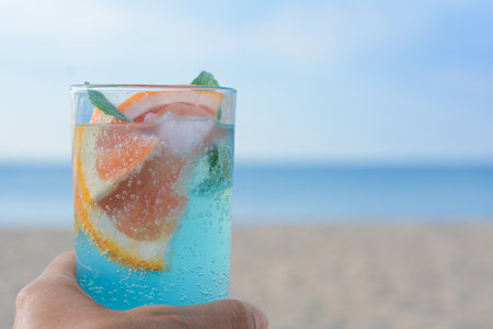 Woman holding glass of refreshing drink with grapefruit and mint at beach, closeup. Space for textの写真素材