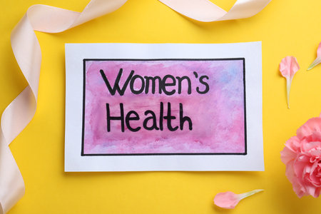 Sheet of paper with words Women's Health, ribbon and carnation flower on yellow background, flat layの写真素材