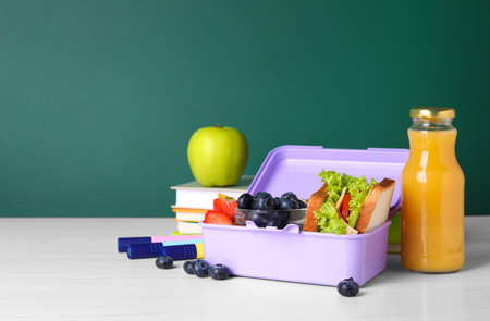 Lunch box with healthy food and different stationery on white wooden table near green chalkboard, space for textの写真素材