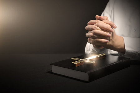 Woman holding hands clasped while praying at table with Bible and cross against dark background, closeup. Space for textの写真素材