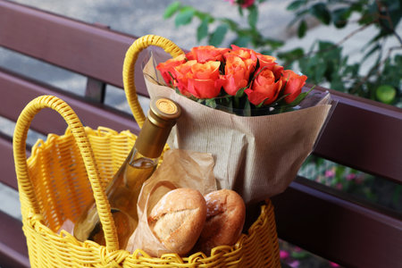 Yellow wicker bag with beautiful roses, bottle of wine and baguettes on bench outdoors, closeupの写真素材