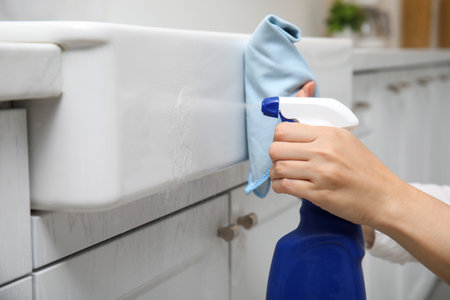 Woman cleaning white sink with rag and detergent in kitchen, closeup