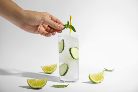 Woman putting mint into glass of tasty fresh cucumber water and sliced lime on white background, closeupの写真素材