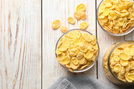 Glass bowls and jar of tasty crispy corn flakes on white wooden table, flat lay. Space for text