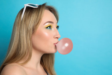 Fashionable young woman with bright makeup blowing bubblegum on light blue background, space for...