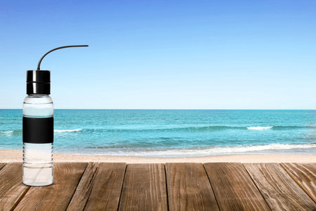Sport bottle with fresh water on wooden table and beautiful view of sandy beach near sea, space for textの写真素材