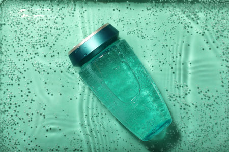 Bottle of hair care cosmetic product in water on turquoise background, top view