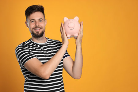 Happy young man with piggy bank on orange background. Space for textの写真素材