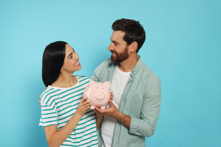 Happy couple with ceramic piggy bank on light blue backgroundの写真素材