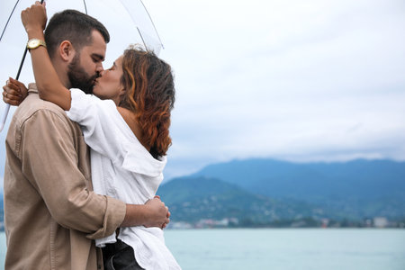 Young couple with umbrella enjoying time together under rain on beach, space for text
