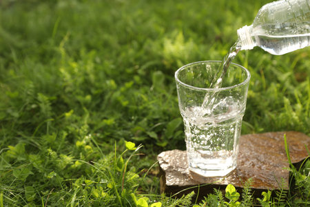 Pouring fresh water from bottle into glass on stone in green grass outdoors. Space for textの写真素材