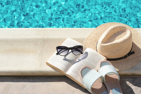 Stylish sunglasses, slippers, straw hat and book at poolside on sunny day. beach accessoriesの写真素材