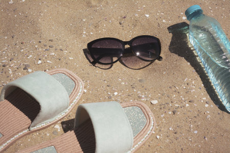 Stylish sunglasses, slippers and bottle of water on sand