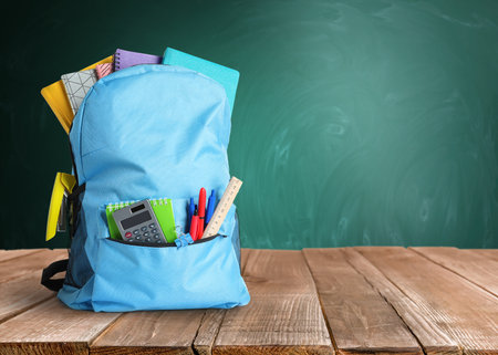 Backpack with school stationery on wooden table near green chalkboard, space for textの写真素材
