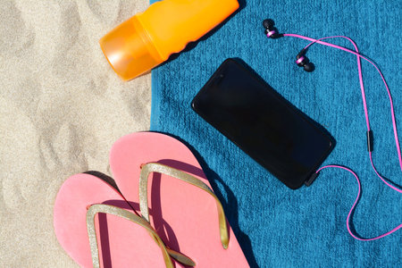 Soft blue beach towel with bottle of sunscreen, smartphone, earphones on sand, flat lay. Space for textの写真素材