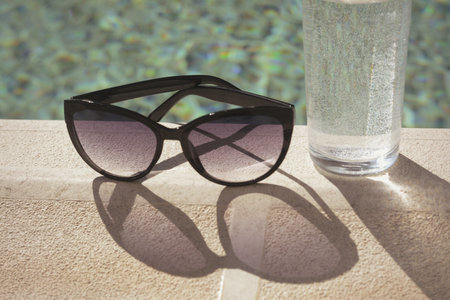 Stylish sunglasses and glass of water near outdoor swimming pool on sunny day, closeupの写真素材