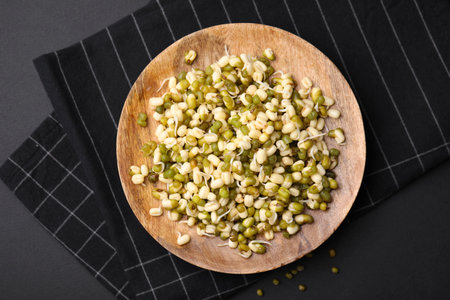 Wooden plate with sprouted green mung beans on black background, flat layの写真素材