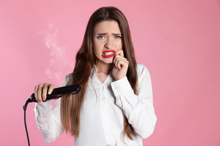 Upset young woman with flattening iron on light pink background. hair damageの写真素材