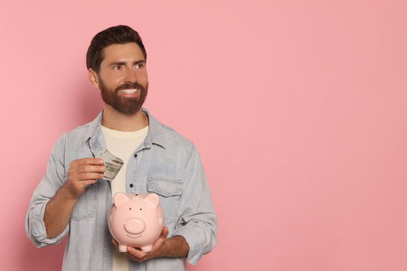 Happy man putting money into piggy bank on pale pink background, space for textの写真素材