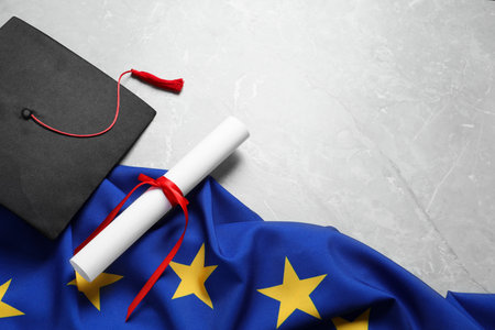 Black graduation cap, diploma and flag of European Union on light gray marble table, above view....