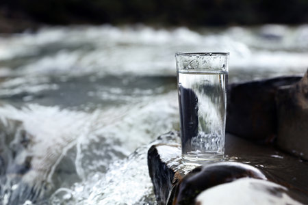 Glass of fresh water on stone near river, space for textの写真素材