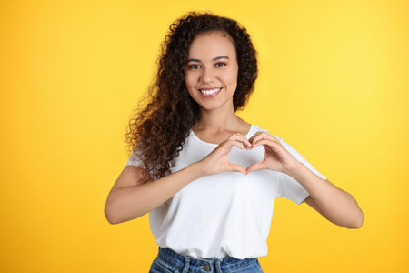 Happy young African-American woman making heart with hands on yellow backgroundの写真素材