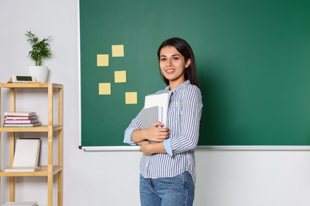 Happy young teacher with books at blackboard in classroom. Space for text