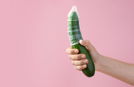 Woman holding cucumber in condom on pink background, closeup and space for text. Safe sex conceptの写真素材