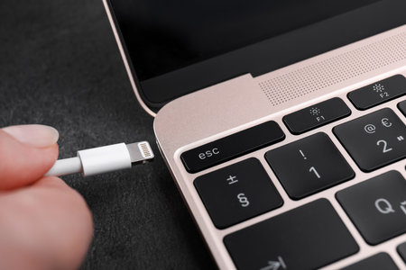 Woman plugging USB cable with lightning connector into laptop port on dark table, closeup