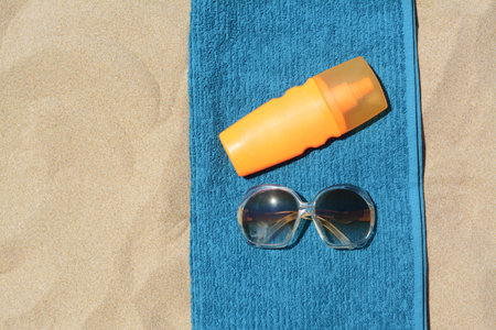 Soft blue beach towel with bottle of sunblock and sunglasses on sand, flat lay. Space for text