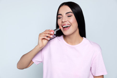 Young woman with beautiful makeup holding nude lipstick on light gray background
