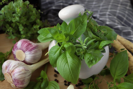 Mortar with different fresh herbs near garlic, horseradish roots and black peppercorns on wooden table, closeupの写真素材