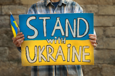 Woman holding poster in colors of national flag and words Stand With Ukraine against brick wall outdoors, closeupの写真素材