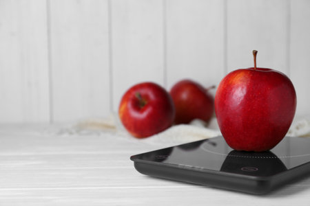 Digital kitchen scale with ripe red apple on white wooden table, space for text