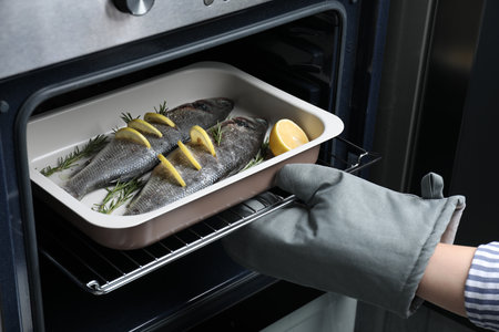Woman putting baking tray with sea bass fish, lemon and rosemary into oven, closeupの写真素材