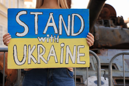 Woman holding poster in colors of national flag with words Stand with Ukraine near broken tank on city street, closeupの写真素材