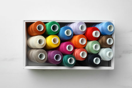 Box with colorful sewing threads on white marble table, top view
