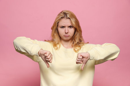 Dissatisfied young woman showing thumbs down on pink backgroundの写真素材