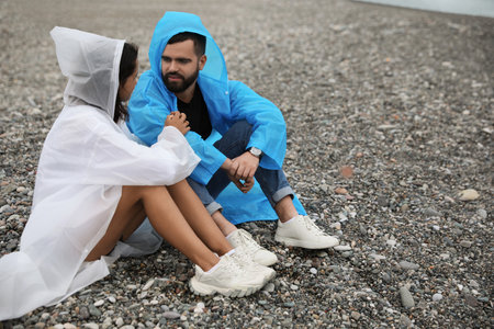 Young couple in raincoats enjoying time together under rain on beach, space for textの写真素材