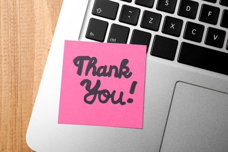 Pink paper note with phrase Thank You and laptop on wooden table, top view