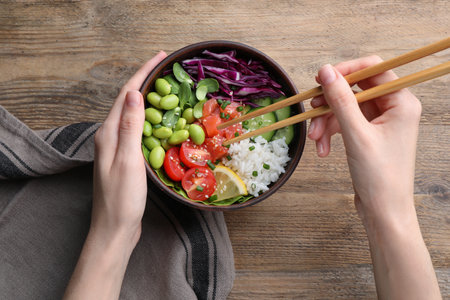 Woman eating poke bowl with salmon, edamame beans and vegetables on wooden table, top viewの写真素材