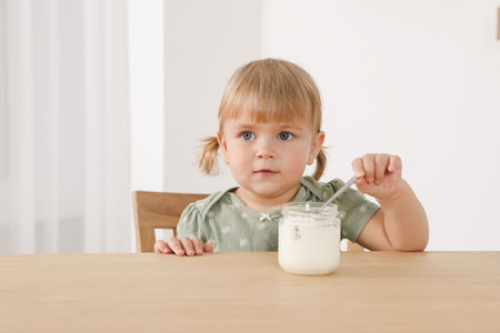 Cute little child eating tasty yogurt with spoon at wooden table indoorsの写真素材