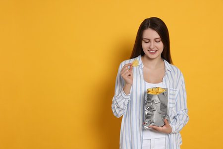 Pretty young woman with bag of tasty potato chips on yellow background. Space for textの写真素材