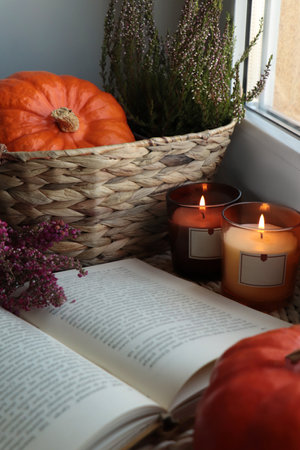 Wicker basket with beautiful heather flowers, pumpkin, burning candles and open book near window indoorsの写真素材