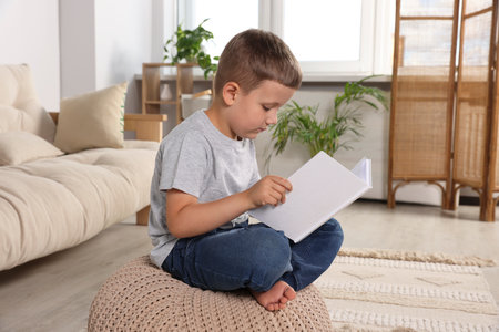 Boy with poor posture reading book on beige pouf in living room. Symptom of scoliosisの写真素材