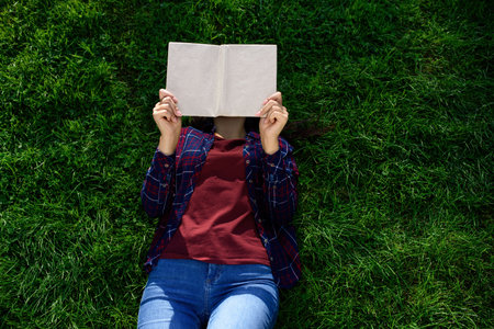 Woman reading book while lying on green grass outdoors, top view