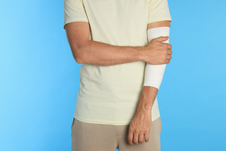 Man with arm wrapped in medical bandage on light blue background, closeupの写真素材