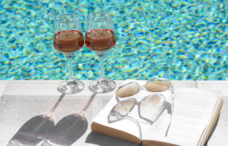 Glasses of tasty rose wine, open book and sunglasses on swimming pool edgeの写真素材